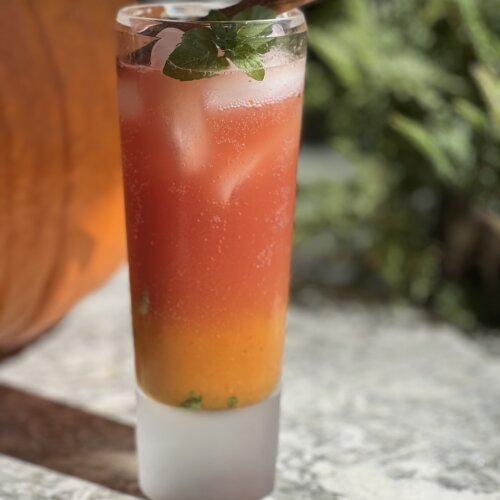 red and orange iced drink in a tall glass with sprig of mint