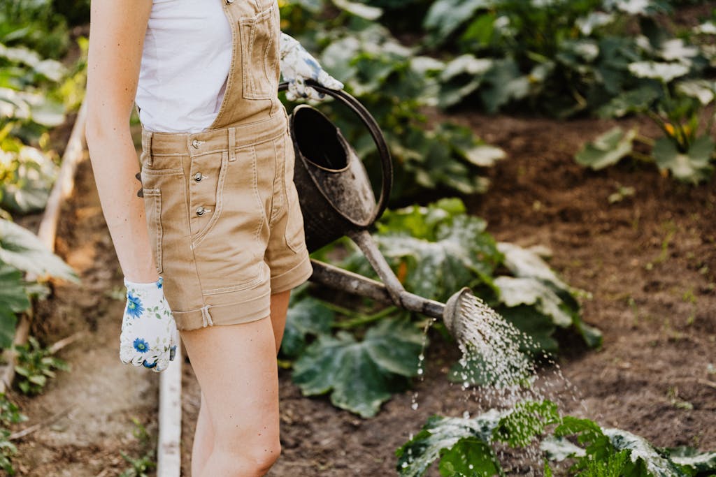 A woman in overalls watering plants in a summer garden with lush greenery.