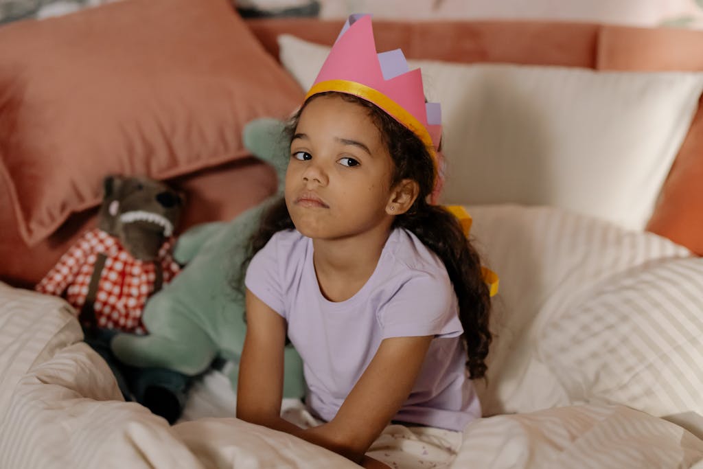 A young girl in a crown sits on a bed with stuffed toys, looking contemplative.